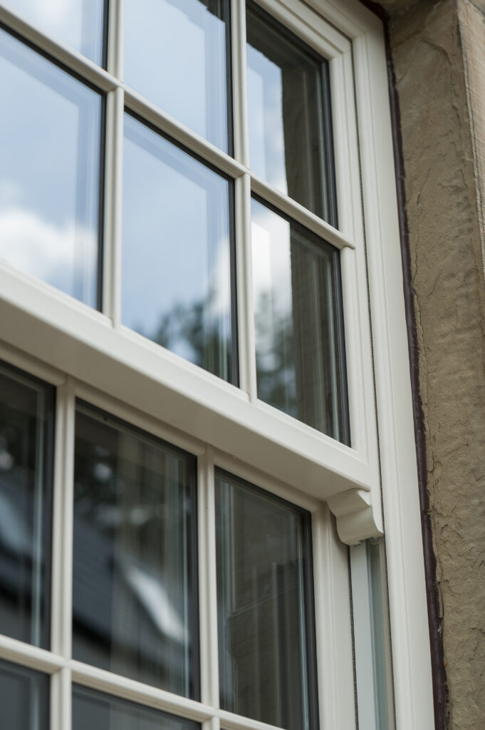 Close-up angled exterior view of white-framed multi-pane windows against a textured wall, with reflections of clouds and soft natural light